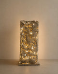 A textured stone pillar lamp with warm white string lights glowing through a rough, rectangular concrete or stone column on a matching base, set against a neutral beige background.