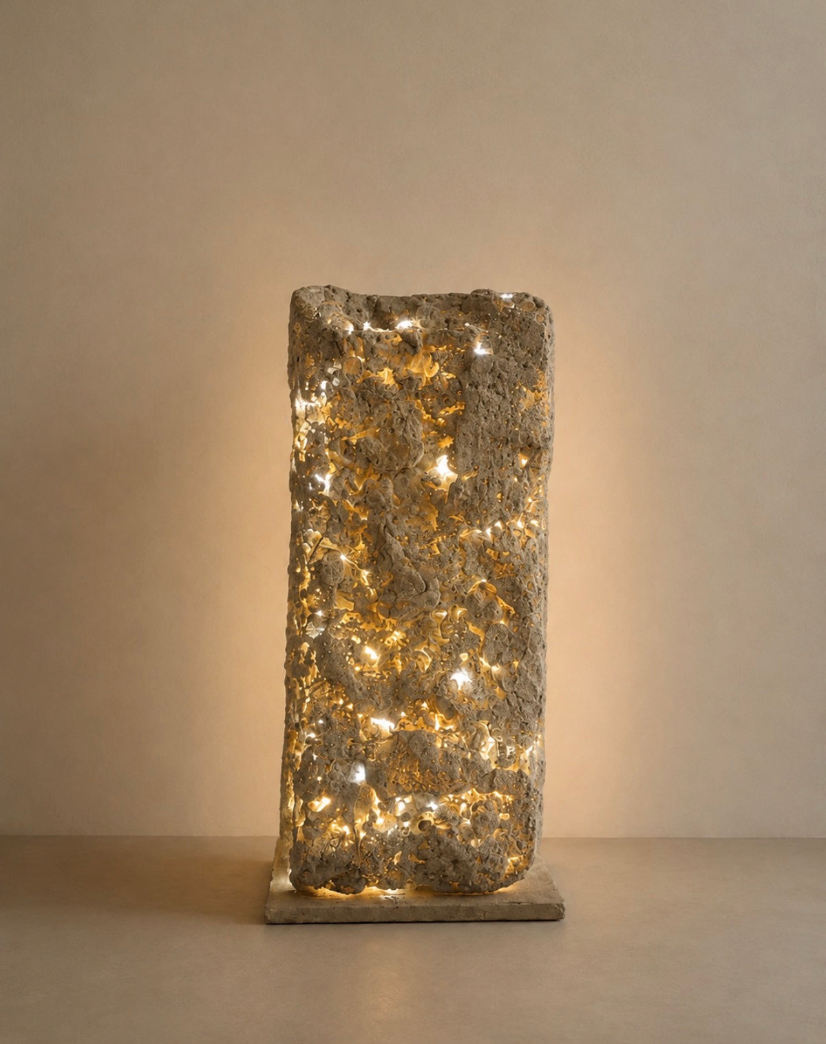 A textured stone pillar lamp with warm white string lights glowing through a rough, rectangular concrete or stone column on a matching base, set against a neutral beige background.