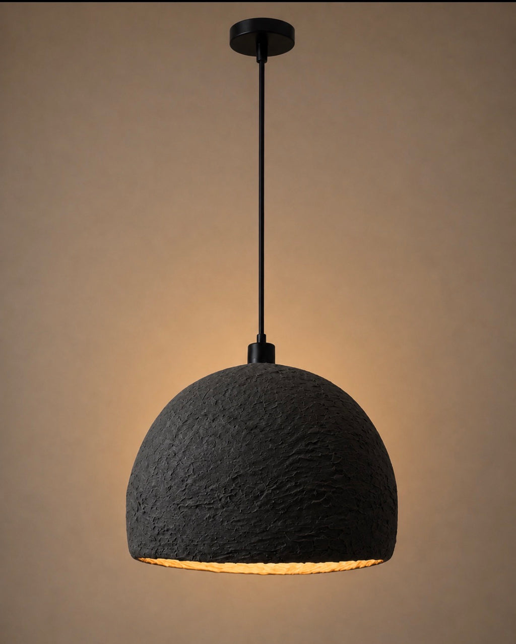 A deeply textured charcoal dome pendant light with a matte black cord and canopy, casting warm amber light against a rich warm beige background.