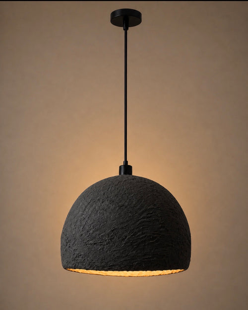 A deeply textured charcoal dome pendant light with a matte black cord and canopy, casting warm amber light against a rich warm beige background.