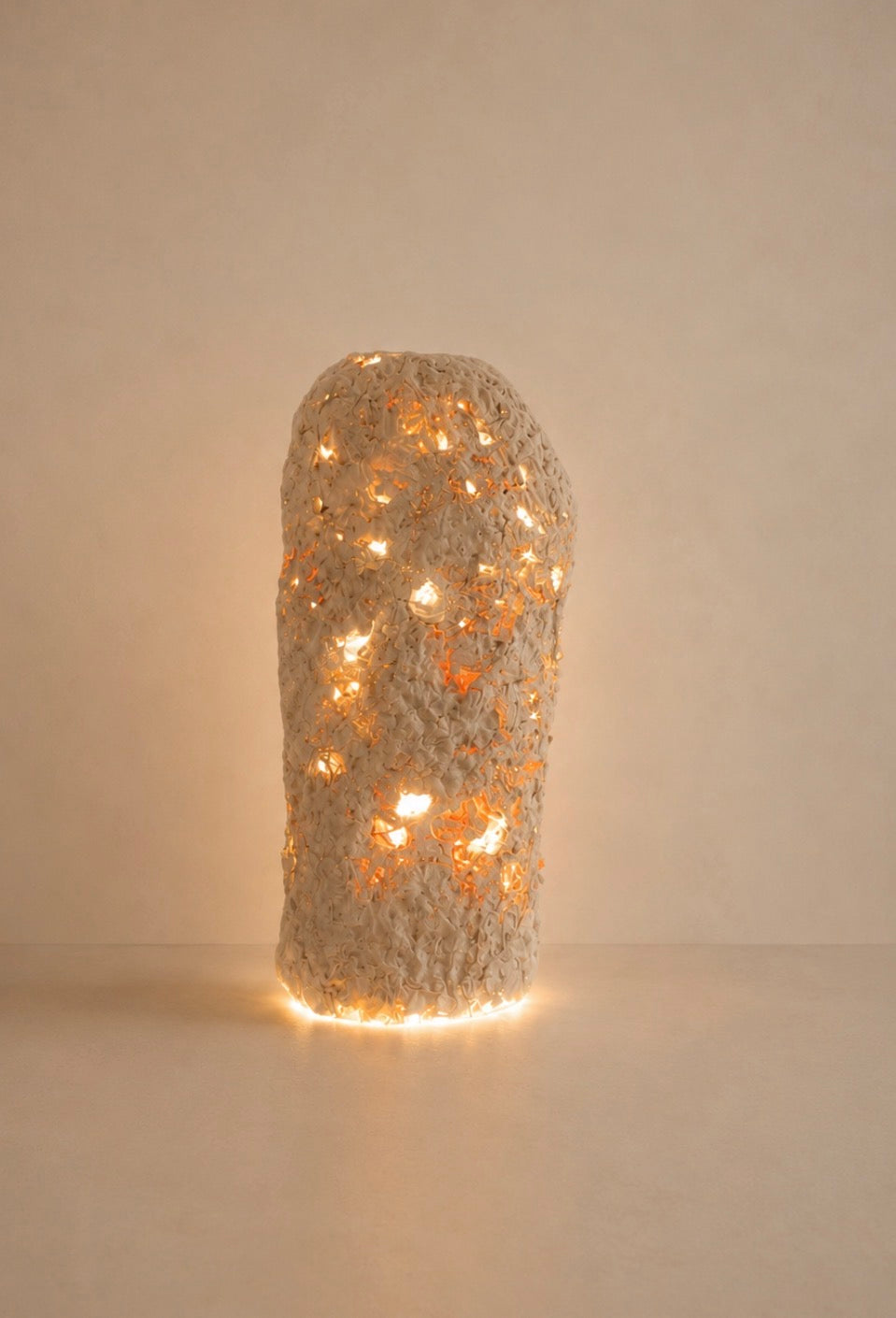 A textured, organic-shaped lamp or sculpture made of a rough, light-colored woven or stone-like material, glowing warmly from internal string lights against a plain beige background.