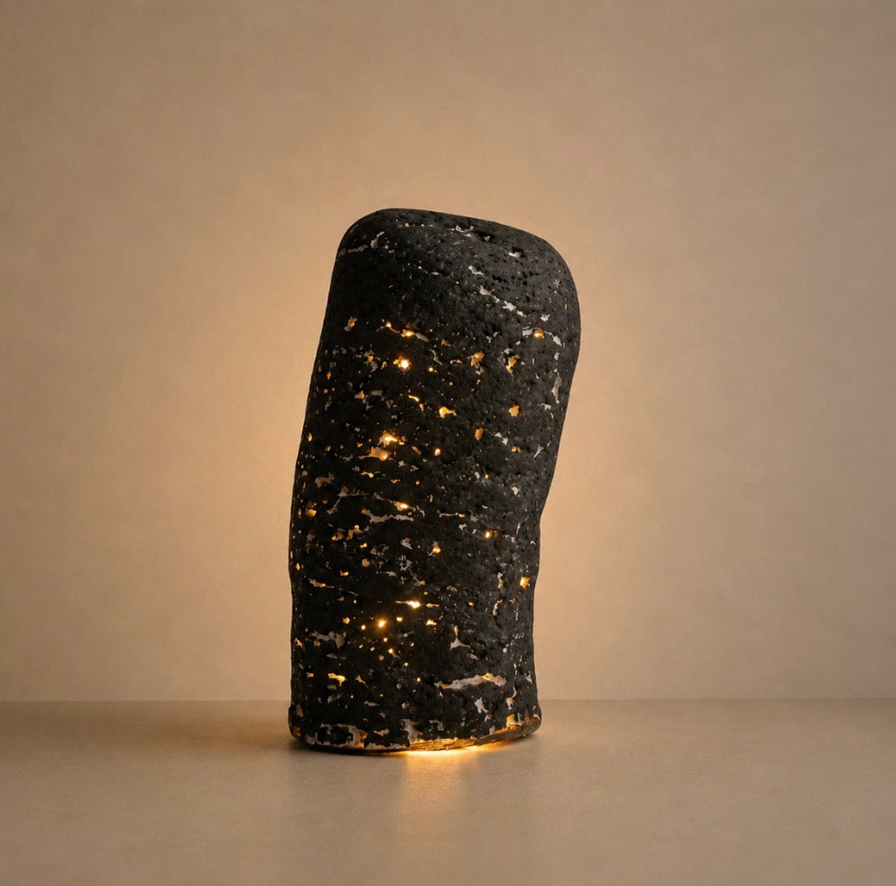 A dark charcoal organic-shaped sculptural lamp with pinpoints of warm golden light piercing through its rough, porous texture, glowing softly against a warm beige background.