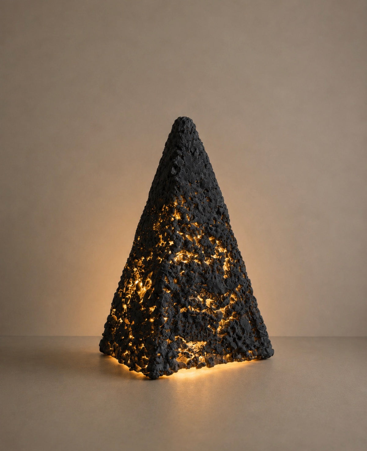 A dark charcoal pyramid-shaped sculptural lamp with warm amber light blazing through its heavily textured surface, casting a dramatic glow onto a neutral background