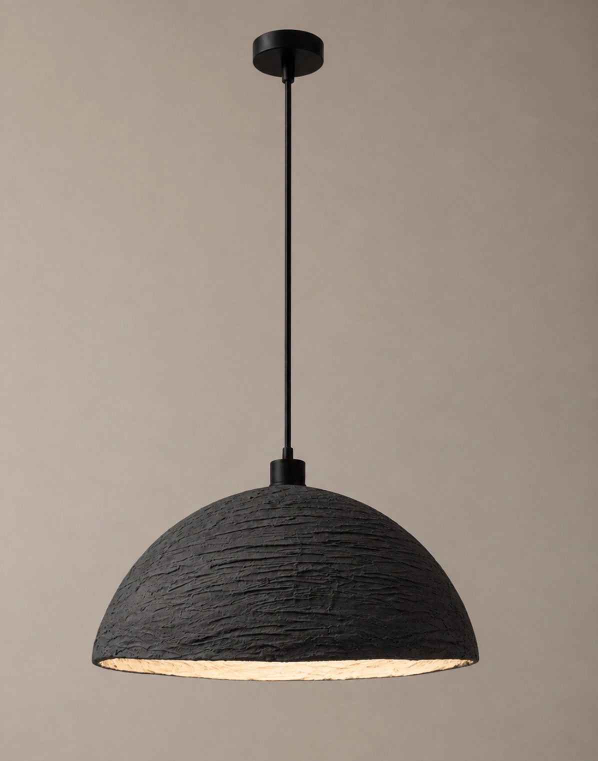 A sculptural pendant light with a wide textured charcoal dome shade and matte black suspension cord, shown against a warm neutral background