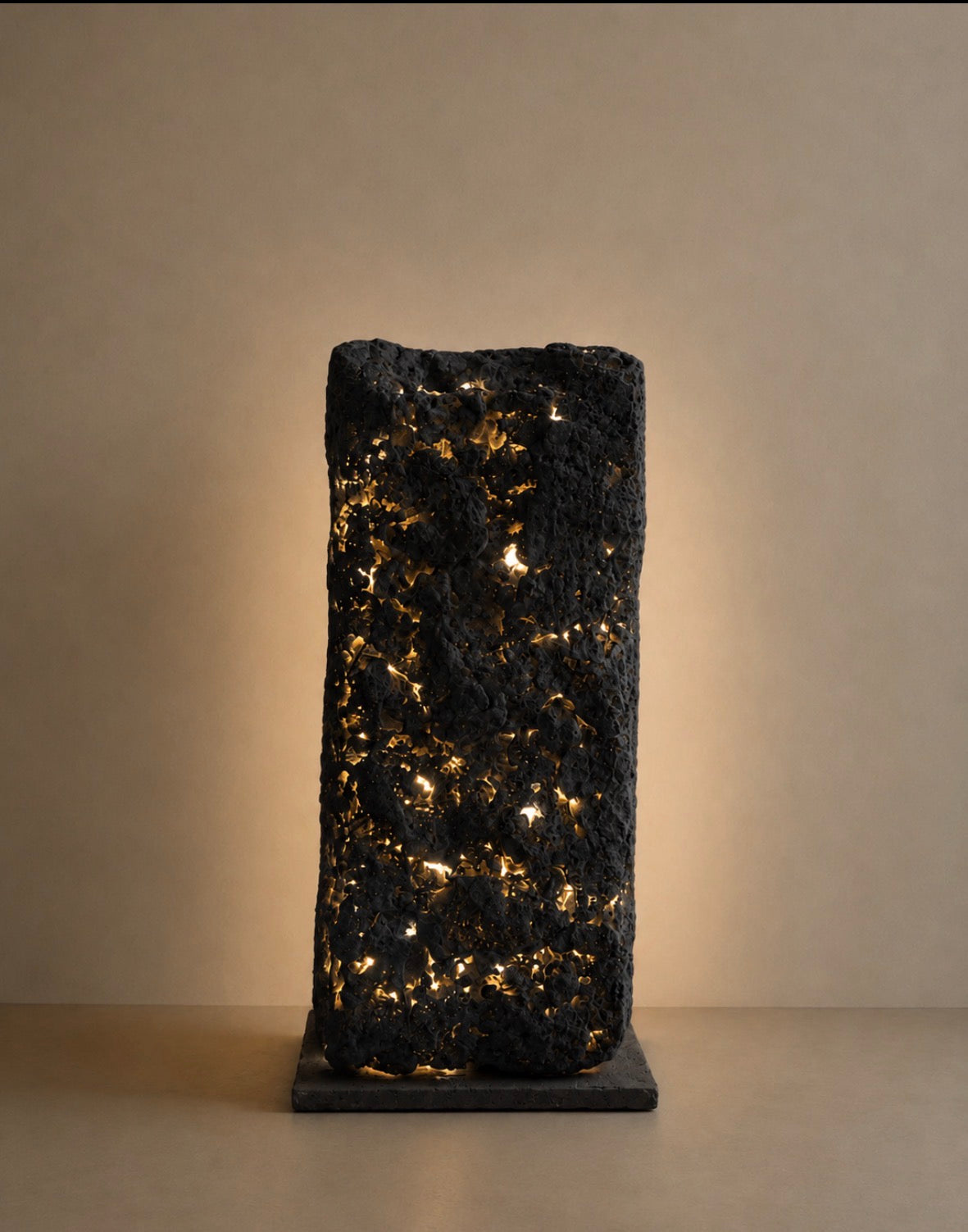 A dark charcoal organic-shaped sculptural lamp with pinpoints of warm golden light piercing through its rough, porous texture, glowing softly against a warm beige background.