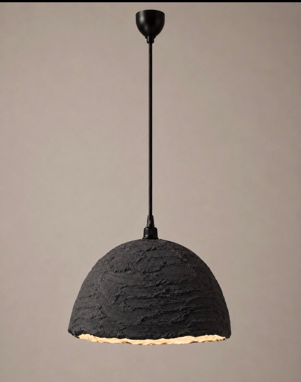 A pendant light with a deeply textured charcoal dome shade and matte black suspension cord and canopy, photographed against a warm neutral background.