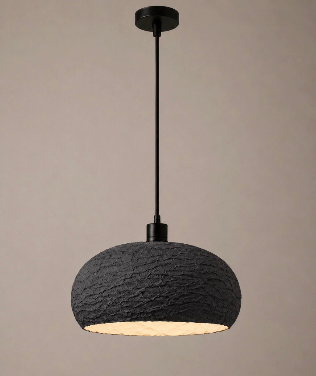 A textured charcoal wide-dome pendant light with a matte black cord and canopy, glowing warmly from within against a soft neutral background
